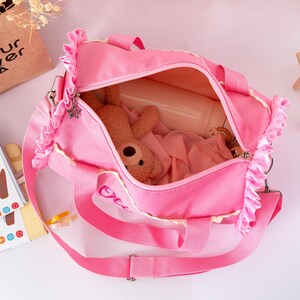 May include: A pink duffel bag with a zipper closure, a shoulder strap, and two handles. The bag has ruffled trim and a star-shaped charm. Inside the bag is a stuffed animal and other items. The bag has the word "Ocean" embroidered on it.