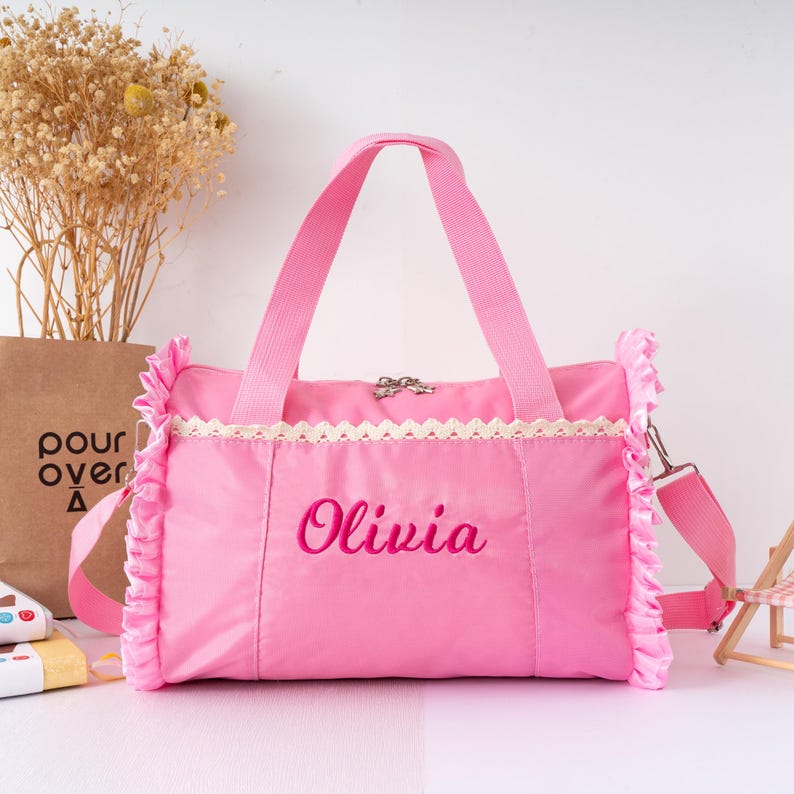 May include: A pink duffel bag with a shoulder strap and handles. The bag has a ruffled trim and a lace detail. The name "Olivia" is embroidered on the front in a cursive font. The bag is sitting on a white surface.