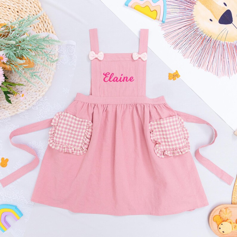 May include: A pink apron with the name "Elaine" embroidered on the front. The apron has two pockets with a pink and white gingham pattern and is decorated with small bows. The apron is designed for children.