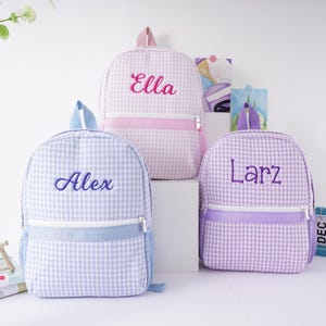 May include: Three personalized gingham backpacks in purple, pink, and blue with the names Alex, Ella, and Larz embroidered on the front.