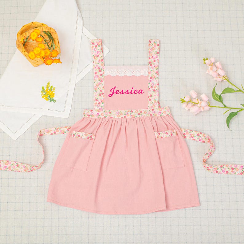 May include: A pink apron with floral patterned straps and pockets. The apron has the name "Jessica" embroidered on the front. The apron has a white lace trim at the top and long floral patterned ties.