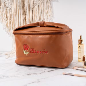 May include: A brown faux leather travel makeup bag with a zipper closure. The bag has a red embroidered rose and the name "Bonnie" on the front.