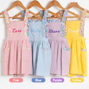 May include: Four children's aprons in pink, blue, purple, and yellow hang on wooden hangers. Each apron has a floral patterned strap and pocket detail. The names "Zara", "Molly", "Diana", and "Coral" are embroidered on the aprons.
