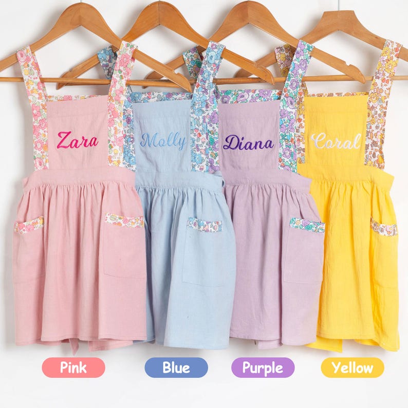 May include: Four children's aprons in pink, blue, purple, and yellow hang on wooden hangers. Each apron has a floral patterned strap and pocket detail. The names "Zara", "Molly", "Diana", and "Coral" are embroidered on the aprons.