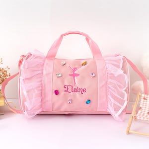 Peut inclure: Sac de sport rose avec un motif de ballerine et le nom "Elaine" brod&eacute; en cursive. Le sac est orn&eacute; d'une bordure &agrave; volants et de bijoux color&eacute;s. Il poss&egrave;de deux poign&eacute;es et une bandouli&egrave;re.