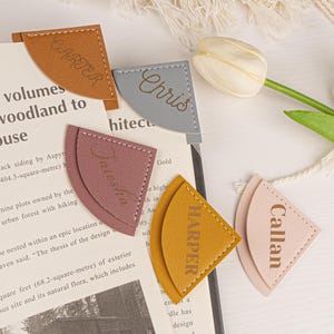 May include: Five personalized leather bookmarks in different colors. The bookmarks are shaped like a triangle with rounded corners and have gold lettering. The names on the bookmarks are Carter, Chris, Aiesha, Harper, and Callan.