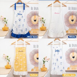 May include: Four aprons with different designs and names are displayed. One is white with blue floral patterns and the name "Vera". Another is white with the name "Monica". A third is yellow with the name "Amelia". The last is white with blue floral patterns and the name "Ali".
