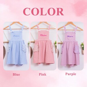 May include: Three children's aprons in blue, pink, and purple, each with a name embroidered on the bib. The aprons have ruffled pockets and bows on the straps. The word "COLOR" is displayed in pink above the aprons.