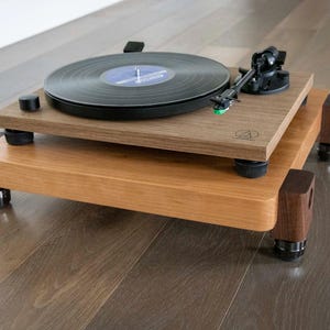 May include: A turntable with a black platter and a wooden base sits on a wooden platform with four legs. The platform has a brown stain and the legs are made of dark wood.