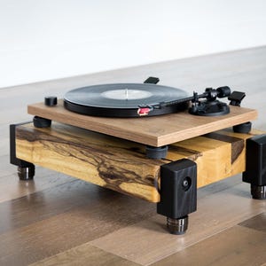 May include: A wooden turntable with a black record, tonearm, and black accents. The turntable sits on a two-tiered wooden base with black corner supports and adjustable feet. The wood grain is visible.