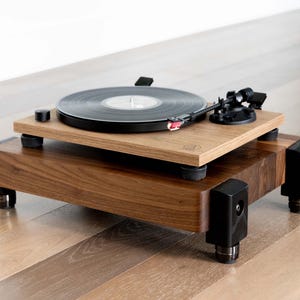 May include: A wooden turntable with a black record playing. The turntable has a light brown top and a darker brown base. The tonearm is black with a red stylus. The turntable sits on a wooden floor.