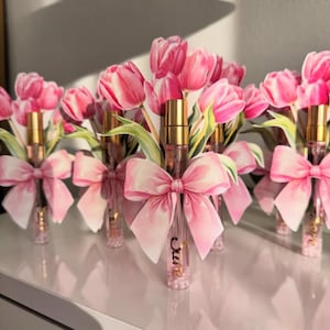 Puede incluir: Varios frascos de perfume con tapas doradas est&aacute;n dispuestos en fila, cada uno adornado con un lazo rosa y un ramo de tulipanes rosas. Los frascos tienen la palabra "cure" escrita.
