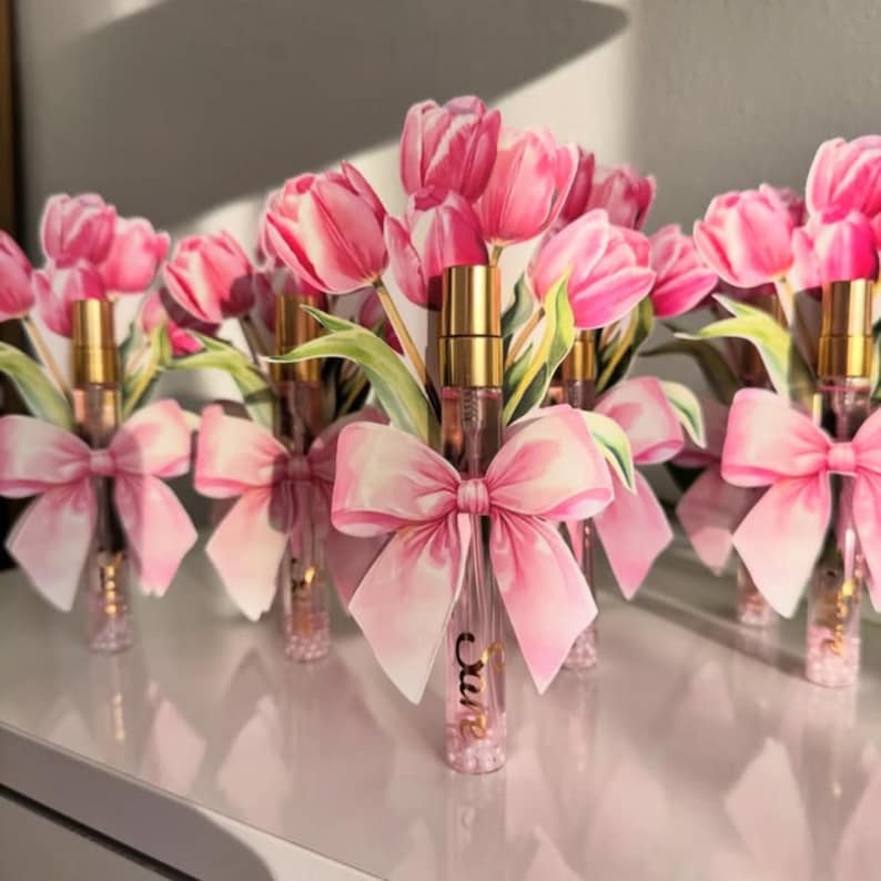 Puede incluir: Varios frascos de perfume con tapas doradas est&aacute;n dispuestos en fila, cada uno adornado con un lazo rosa y un ramo de tulipanes rosas. Los frascos tienen la palabra "cure" escrita.