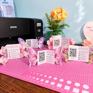 Könnte beinhalten: Schmetterlingsförmige Visitenkarten in Rosa und Lila mit QR-Codes und Kontaktdaten liegen auf einer rosa Schneidematte. Ein Epson-Drucker und künstliche Blumen befinden sich im Hintergrund.