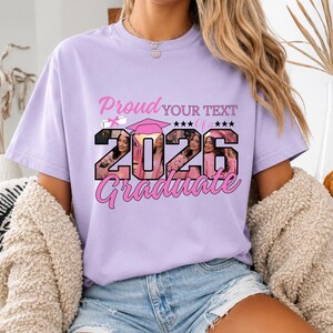 May include: A light purple t-shirt with the text "Proud YOUR TEXT of a 2026 Graduate" in pink. The numbers 2026 are filled with a photo of a person wearing a graduation cap. The shirt has short sleeves.