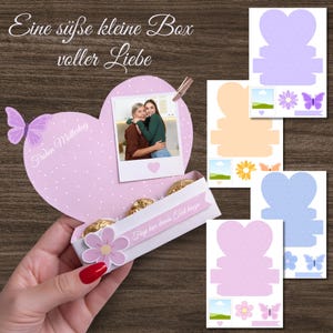 May include: A heart-shaped gift box with a photo of two people hugging, a pink flower, and a butterfly. The box is filled with gold-wrapped chocolates. The text on the box reads "Eine süße kleine Box voller Liebe" and "Füge hier deinen Text hinzu."