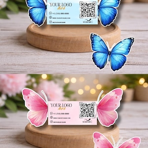 Może przedstawiać: Wizytówki z motywami motyli. Jedna karta jest jasnoniebieska z niebieskimi motylami, druga różowa z różowymi motylami. Każda karta zawiera logo, dane kontaktowe i kod QR. Karty są prezentowane na drewnianym stojaku.