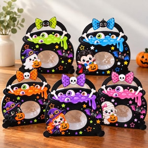 May include: Five black cardboard Halloween treat bags shaped like cauldrons, each featuring a clear circular window, a colorful bow, and illustrations of ghosts, vampires, cats, or mummies with eyeballs and bones in neon-colored bubbling liquid.