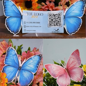 Puede incluir: Decoraciones con forma de mariposa en azul y rosa, dispuestas con flores. También hay una tarjeta de visita con el texto "YOU LOGO here", un número de teléfono, un sitio web y un nombre de Instagram. La tarjeta incluye un código QR y "SCAN ME".