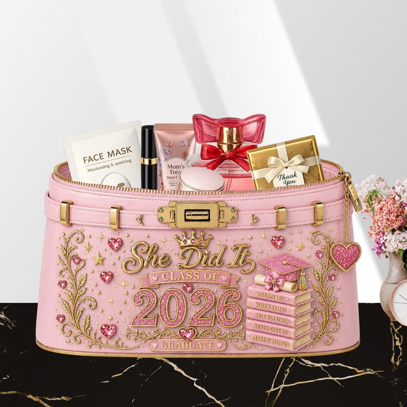 May include: A pink graduation gift box filled with beauty products. The box is decorated with gold accents, hearts, and the text "She Did It Class of 2026 Graduate." Inside are a face mask, lipstick, perfume, and a small gift box.