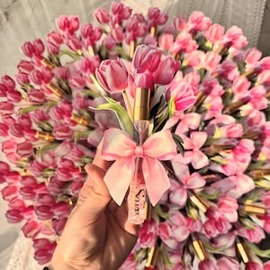 Puede incluir: Un frasco de perfume rosa con tapa dorada y lazo rosa se sostiene frente a un ramo de tulipanes rosas. La botella tiene la palabra "Love" impresa. Los tulipanes tienen detalles blancos.