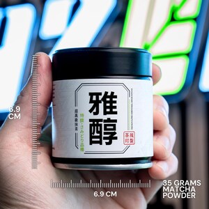 Puede incluir: Una lata cil&iacute;ndrica negra de polvo de matcha con una etiqueta blanca que presenta los caracteres japoneses 雅醇. La etiqueta indica 35 gramos de polvo de matcha y una altura de 6,9 cm, seg&uacute;n se muestra en una escala de medici&oacute;n visible.
