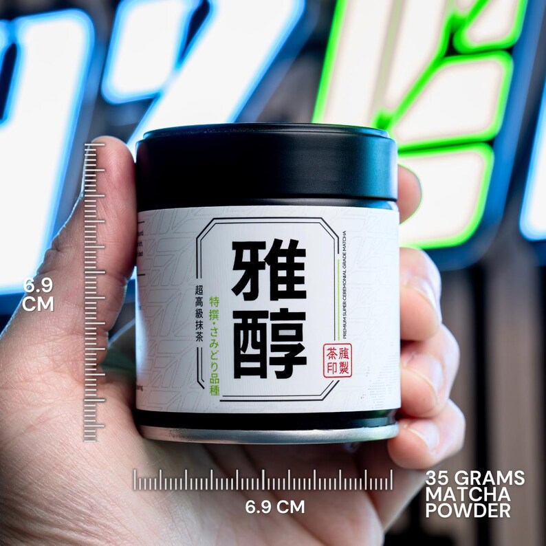 Puede incluir: Una lata cil&iacute;ndrica negra de polvo de matcha con una etiqueta blanca que presenta los caracteres japoneses 雅醇. La etiqueta indica 35 gramos de polvo de matcha y una altura de 6,9 cm, seg&uacute;n se muestra en una escala de medici&oacute;n visible.