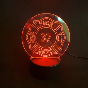 May include: A glowing red LED night light featuring a firefighter's Maltese cross emblem. The design includes the words "FIRE DEPT." and the number "37" in a circular format. The light sits on a black base.
