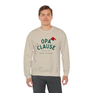Peut inclure: Un sweat-shirt beige avec un motif vert et rouge qui dit "OPA CLAUSE" avec un chapeau de Père Noël au-dessus du texte et "MERRY & BRIGHT" en dessous du texte.