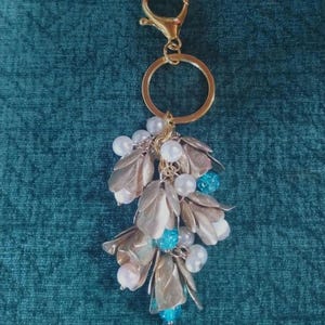 May include: A gold-toned metal keychain featuring a lobster clasp and a circular ring. The charm cluster consists of multiple metallic petal-shaped components in a soft champagne hue, interspersed with round white faux pearls and bright blue beads.