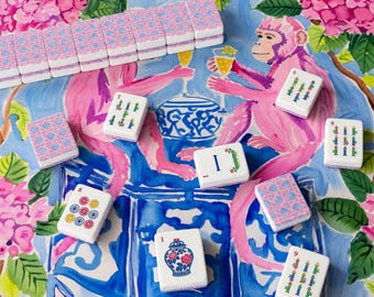May include: A Mahjong set featuring white tiles with colorful illustrations of monkeys, floral patterns, and blue and white ginger jars. The tiles are arranged on a surface decorated with a vibrant painted scene of pink monkeys and hydrangea flowers.
