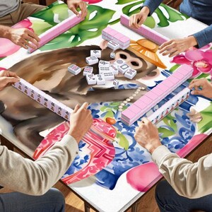 May include: A group of people playing Mahjong. The game is set on a table with a colorful floral and monkey design. Pink tiles and racks are visible, with players holding their tiles. The table is surrounded by hands.