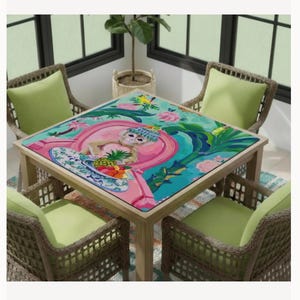 May include: A square wooden table with a colorful, tropical-themed tabletop featuring a monkey, flowers, and fruit. The table is surrounded by four wicker chairs with green cushions, creating a vibrant outdoor dining set.