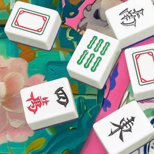 Op de afbeelding: Een verzameling witte Mahjong-tegels met verschillende symbolen en markeringen in rood, groen en zwart. De tegels liggen verspreid op een kleurrijk bloemenpatroon, wat een levendige compositie cre&euml;ert. Klassieke bordspelstukken.