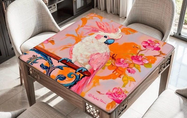May include: A square wooden table with a vibrant, artistic design. The tabletop features a painting of a pink and white cockatoo perched on a branch with pink flowers against an orange and pink background. The table has a decorative wooden frame.