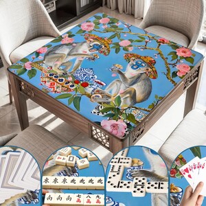 K&ouml;nnte beinhalten: Ein Spieltisch aus Holz mit einer blauen Oberfl&auml;che, die ein skurriles Design von Affen, Blumen und &Auml;sten zeigt. Der Tisch ist von St&uuml;hlen umgeben und enth&auml;lt Nahaufnahmen von Spielsteinen wie Karten, Dominosteinen und Mahjong-Steinen. Der Tisch misst ca. 91 cm im Quadrat.