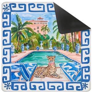 May include: A rectangular mousepad featuring a watercolor painting of a cheetah lounging by a pool. The scene includes a pink building, palm trees, and blue and white decorative elements. The border is white with a blue Greek key design.