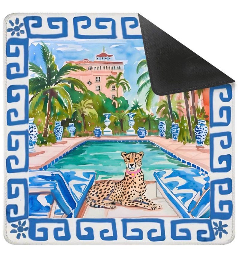 May include: A rectangular mousepad featuring a watercolor painting of a cheetah lounging by a pool. The scene includes a pink building, palm trees, and blue and white decorative elements. The border is white with a blue Greek key design.