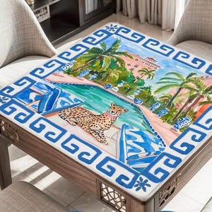 May include: Square wooden table with a colorful, tropical-themed top. The artwork depicts a cheetah by a pool, palm trees, and a resort. The border is a blue and white Greek key design. The table is surrounded by two chairs.