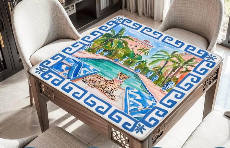 May include: Square wooden table with a colorful, tropical-themed top. The artwork depicts a cheetah by a pool, palm trees, and a resort. The border is a blue and white Greek key design. The table is surrounded by two chairs.