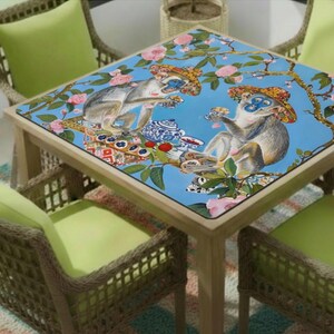 K&ouml;nnte beinhalten: Quadratischer Holztisch mit blauer Tischplatte, verziert mit einer verspielten Illustration von Affen mit H&uuml;ten, die Tee genie&szlig;en. Der Tisch ist von Korbst&uuml;hlen mit gr&uuml;nen Kissen umgeben.