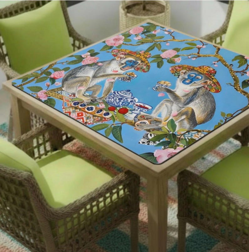 K&ouml;nnte beinhalten: Quadratischer Holztisch mit blauer Tischplatte, verziert mit einer verspielten Illustration von Affen mit H&uuml;ten, die Tee genie&szlig;en. Der Tisch ist von Korbst&uuml;hlen mit gr&uuml;nen Kissen umgeben.