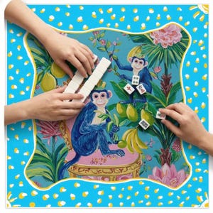 May include: A vibrant Mahjong game set with a playful design. The game board has a blue background with a monkey illustration, a yellow border, and white polka dots. Hands are shown playing the game with white tiles.
