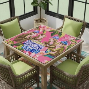 May include: A square wooden table with a pink surface featuring a monkey design. The table is surrounded by four wicker chairs with green cushions. The monkey design includes monkeys wearing hats and playing cards.