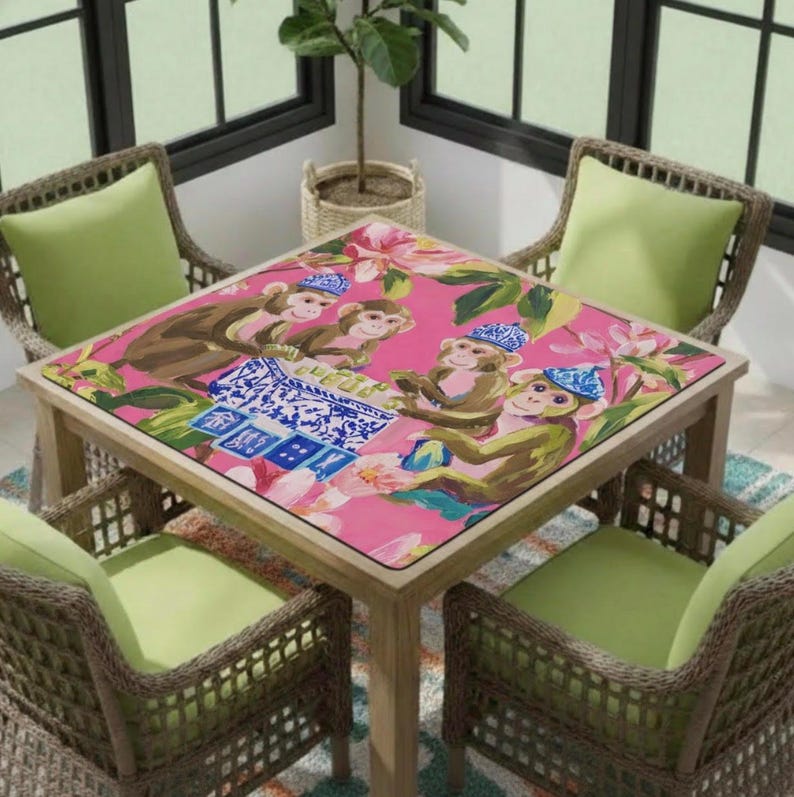 May include: A square wooden table with a pink surface featuring a monkey design. The table is surrounded by four wicker chairs with green cushions. The monkey design includes monkeys wearing hats and playing cards.