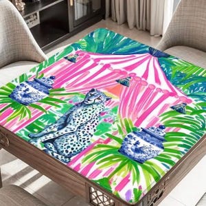 Puede incluir: Un mantel cuadrado con un patr&oacute;n vibrante de un guepardo, tarros de jengibre azules y blancos, y hojas de palma verdes sobre un fondo de rayas verticales de color rosa brillante y blanco. El dise&ntilde;o es colorido, detallado y gr&aacute;fico.