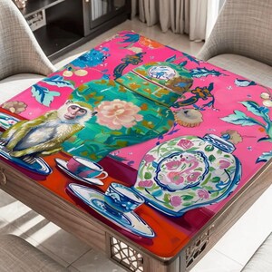 Op de afbeelding: Vierkante houten tafel met een felroze blad met een speels ontwerp. Het ontwerp omvat een aap, kopjes en decoratieve potten met bloemmotieven. De tafel wordt omringd door twee stoelen.
