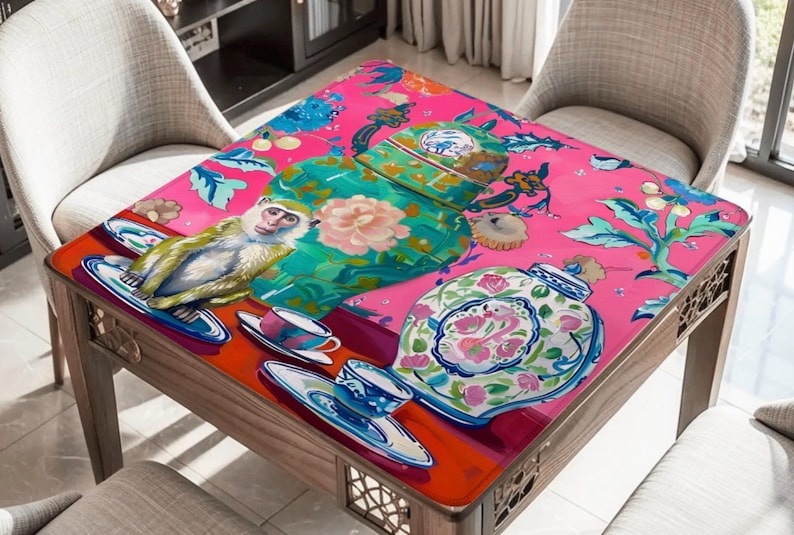 Op de afbeelding: Vierkante houten tafel met een felroze blad met een speels ontwerp. Het ontwerp omvat een aap, kopjes en decoratieve potten met bloemmotieven. De tafel wordt omringd door twee stoelen.