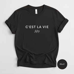May include: Black t-shirt with rolled sleeves. The shirt features the phrase "C'EST LA VIE" above the word "fifty" in a white, sans-serif font. A black circle with the word "black" is in the lower right corner.