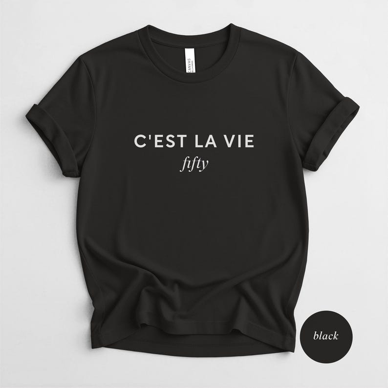 May include: Black t-shirt with rolled sleeves. The shirt features the phrase "C'EST LA VIE" above the word "fifty" in a white, sans-serif font. A black circle with the word "black" is in the lower right corner.